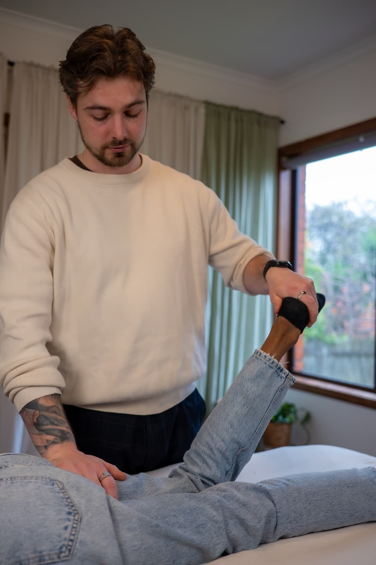 Vildan Alihodzic holds a client's raised foot and steadies her hip for a leg muscle test, clinic window behind.